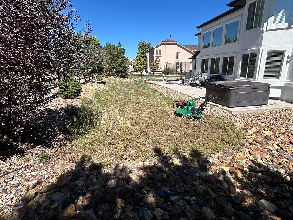 A backyard with patchy grass, a green lawn aerator, a hot tub on a concrete patio, and rocky landscaping, bordered by trees and a fence, with houses in the background under a clear blue sky.