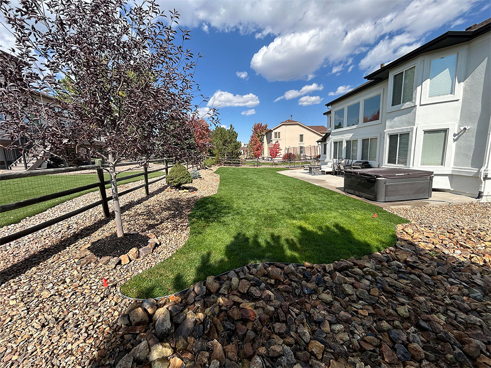A backyard with a curved green lawn bordered by rocks, small trees, and shrubs. A white two-story house with a patio, hot tub, and outdoor furniture is on the right under a partly cloudy sky.