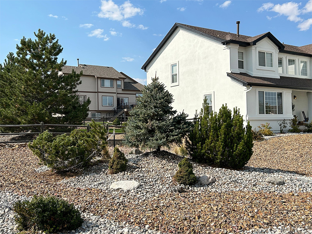 A suburban house with a white exterior and a gravel-covered front yard featuring small evergreen trees and shrubs under a blue sky with scattered clouds. Another house and trees are visible in the background.