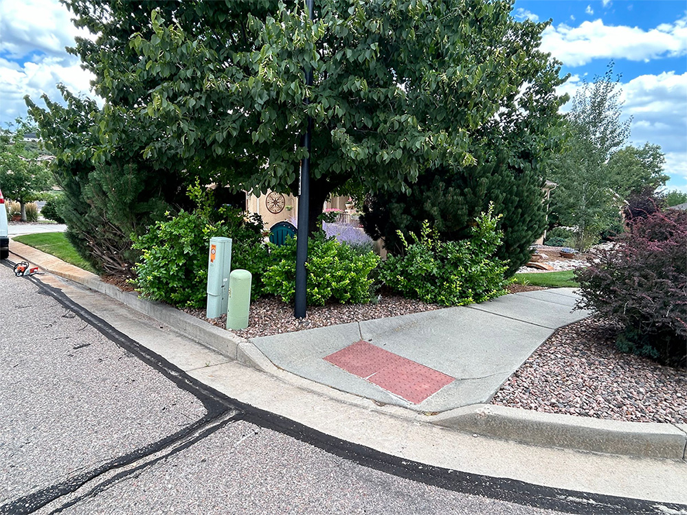 A sidewalk curb ramp with a red tactile paving leads from the street to a path surrounded by green bushes and trees, next to a utility box and a sloped curb on a partly cloudy day.