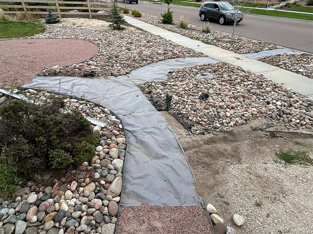 A residential front yard with a partially finished rock landscape, visible landscaping fabric, and areas of exposed dirt. A sidewalk, a car, and a street are in the background.