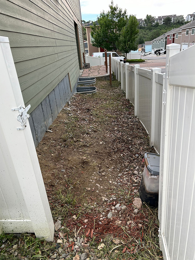 A narrow, rocky strip of bare ground runs alongside a green house, bordered by a white vinyl fence. A few utility boxes and window wells are visible, with trees and parked cars in the background.