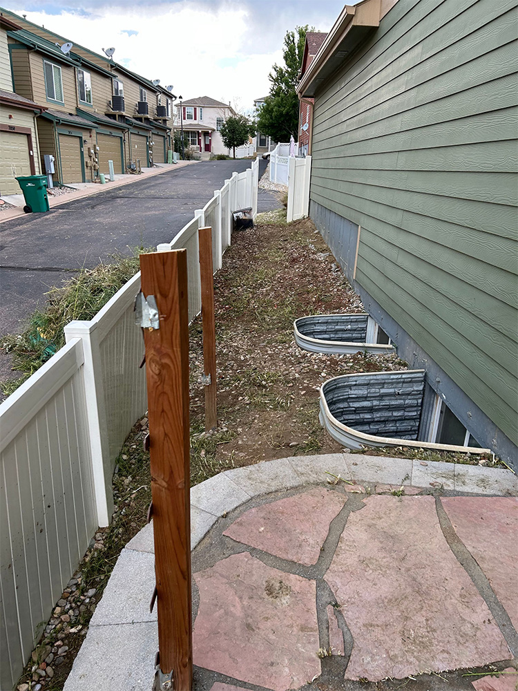 A narrow side yard beside a green house with stone steps, white fence posts, and window wells; some fence panels are missing and the area has patchy grass and gravel.