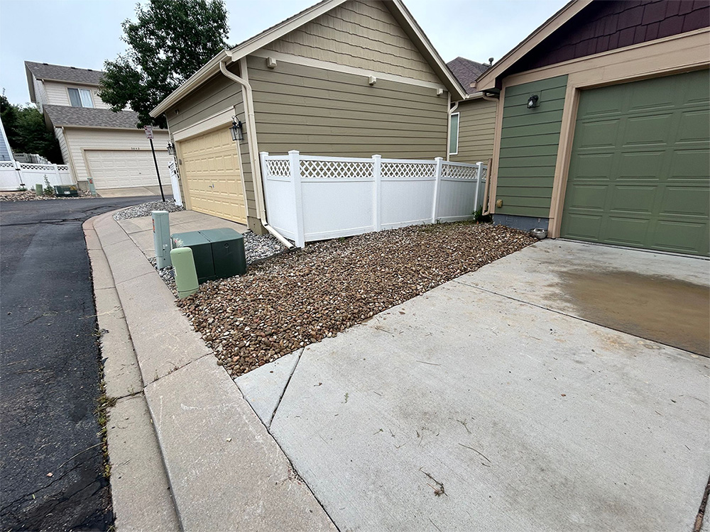 A small gravel yard bordered by a white fence next to a tan garage, with a concrete driveway and nearby green utility boxes along a residential street.