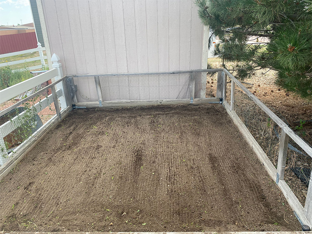 A rectangular raised garden bed filled with freshly tilled soil sits beside a light-colored shed and a pine tree, surrounded by mesh fencing and white railings.
