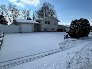 A light gray split-level house with a snowy front yard and driveway, bare trees in the background, and a "For Sale" sign near the curb on a clear winter day—perfect for before and after transformation photos.