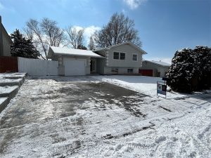 A light gray split-level house with a single-car garage and a snow-covered driveway. Bare trees stand in the background, and there’s a real estate sign in the snow near the driveway entrance.