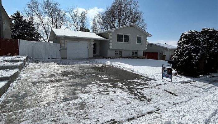 A light gray split-level house with a single-car garage and a snow-covered driveway. Bare trees stand in the background, and there’s a real estate sign in the snow near the driveway entrance.