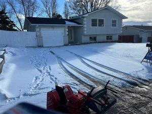A snow-covered driveway leads to a light gray house with tracks and footprints in the snow. A red snowblower sits in the foreground, and bare trees stand behind the house under a partly cloudy sky.