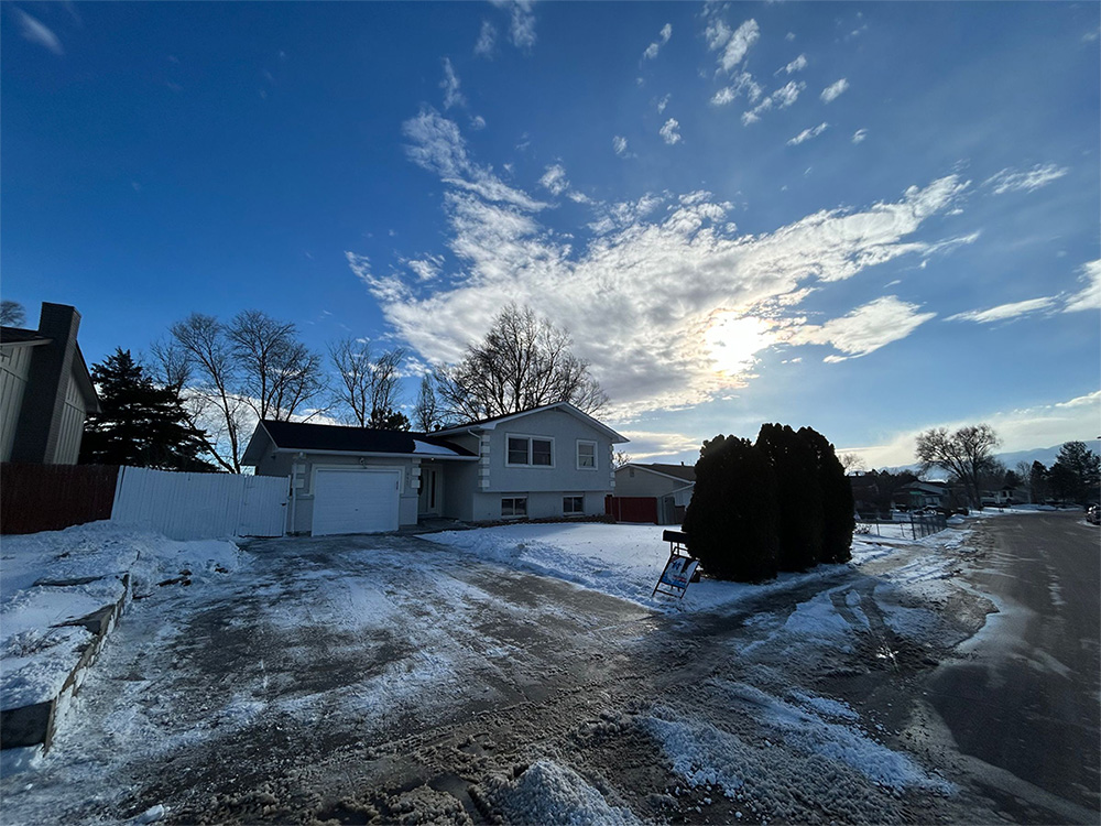 A suburban house in winter with snow on the ground, a cleared driveway, and bare trees. The sun is partially obscured by clouds in a bright blue sky. Shrubs stand near the driveway, and a street runs alongside.