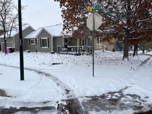 A snow-covered residential street corner with a stop sign and houses. Brown leaves cling to trees, and tire tracks are visible in the snow. A sidewalk and yard are partially covered by the fresh snow.