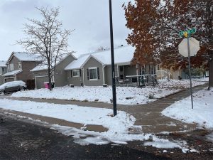 A suburban house on a corner lot is surrounded by snow, with patches of grass and fallen autumn leaves visible. A stop sign and street sign are at the intersection. Cars and bare trees are in the background.