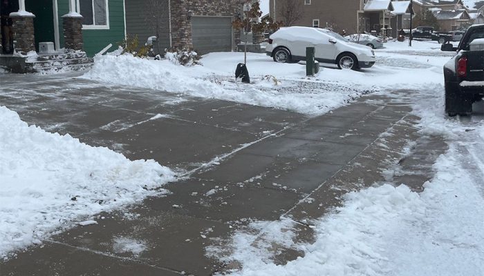A residential neighborhood with snow-covered lawns and driveways, some areas shoveled clear. A snow shovel and snow-covered cars are visible; houses line the street under a cloudy sky.