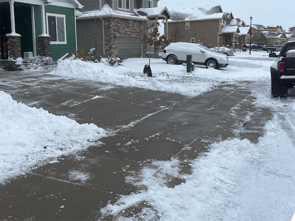 A residential neighborhood with snow-covered lawns and driveways, some areas shoveled clear. A snow shovel and snow-covered cars are visible; houses line the street under a cloudy sky.